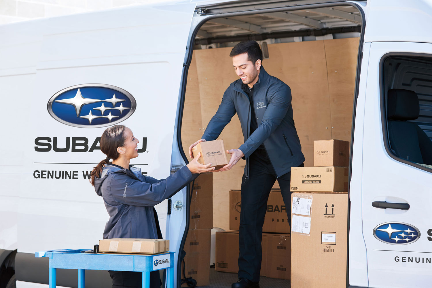 Two employees in blue Subaru jackets exchange a box at the rear of a white van with Subaru branding.