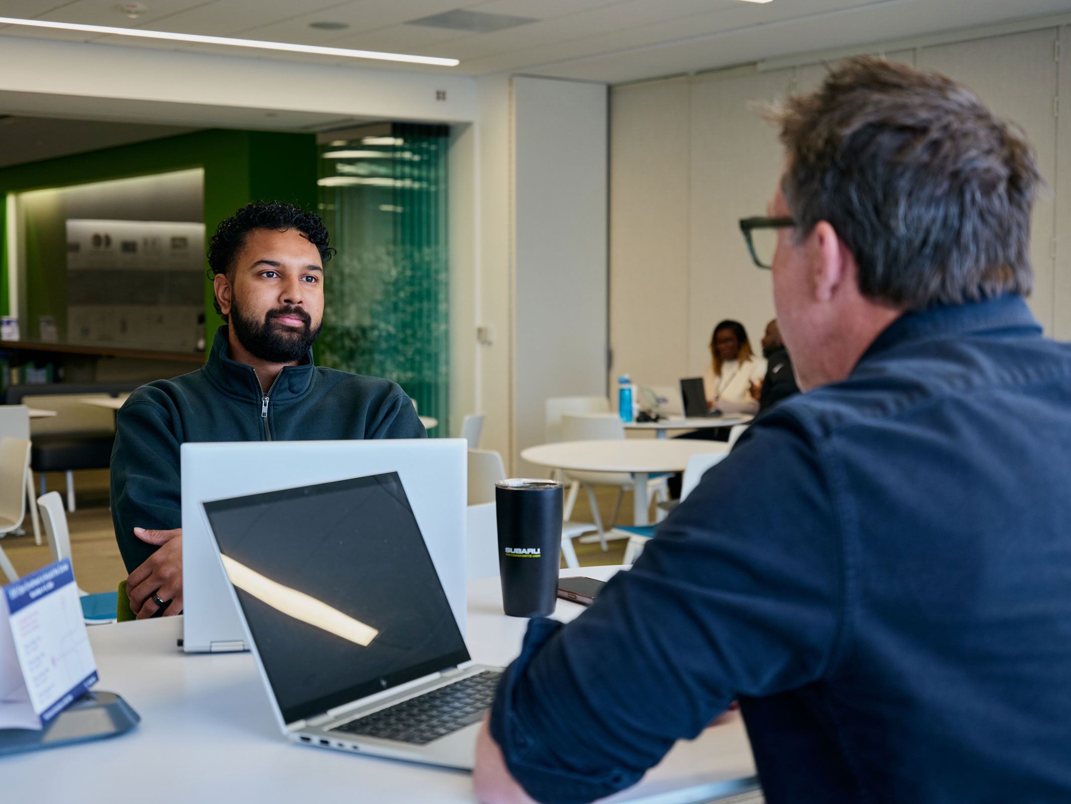 Two people sit at a table in a modern office, discussing. Laptops and a coffee cup are on the table.
