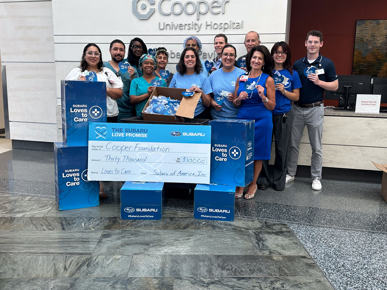 A group of smiling people stand in a hospital lobby, holding a large check for $30,000 from Subaru to the Cooper Foundation. Boxes labeled 