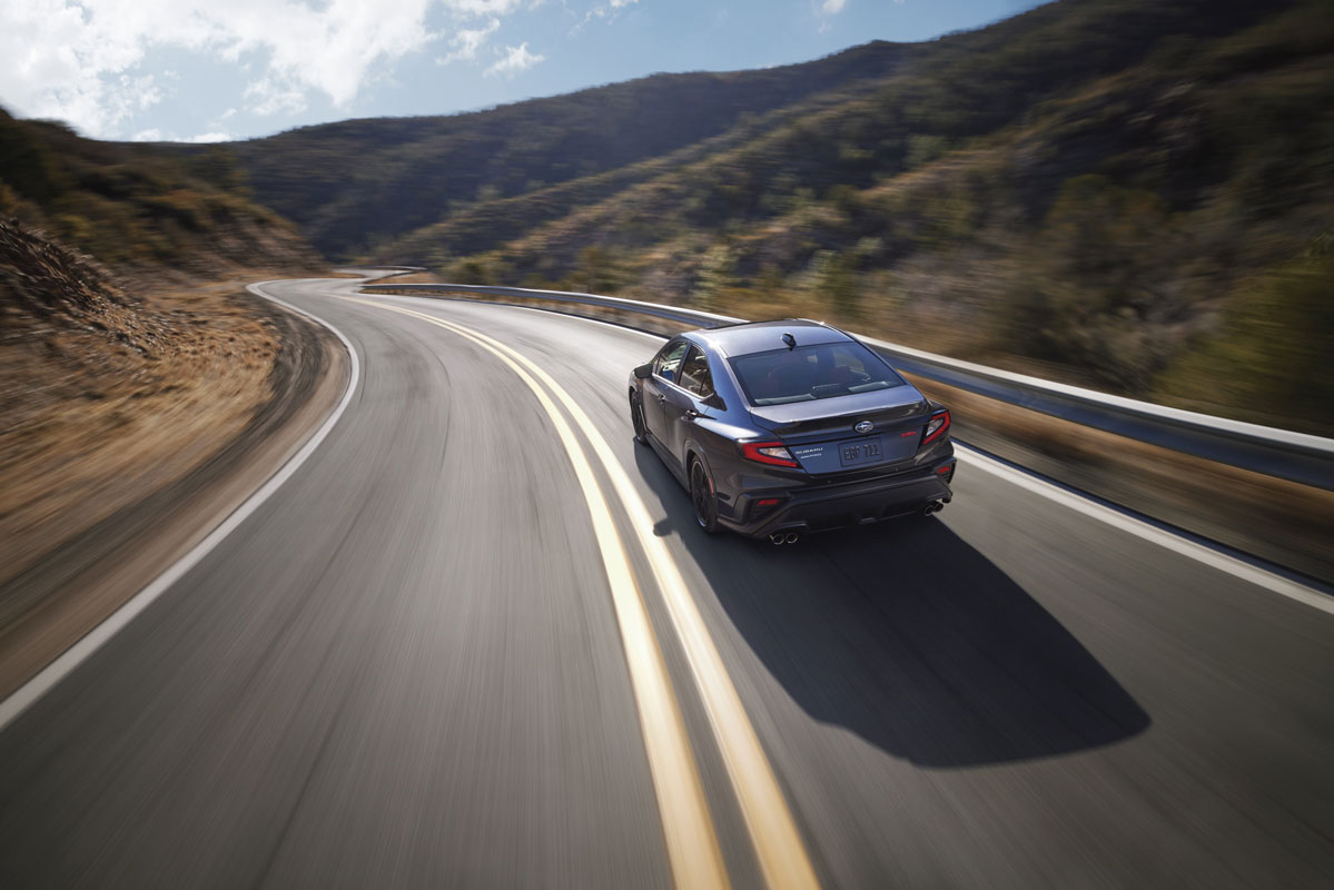 A sleek black Subaru WRX speeds down a winding mountain road under a clear blue sky.