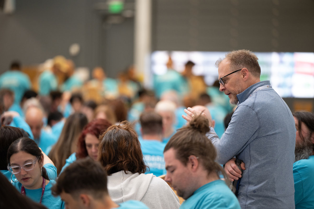 Man in blue jacket speaking to a seated crowd, mostly wearing turquoise Subaru t-shirts, in a busy, focused indoor setting.