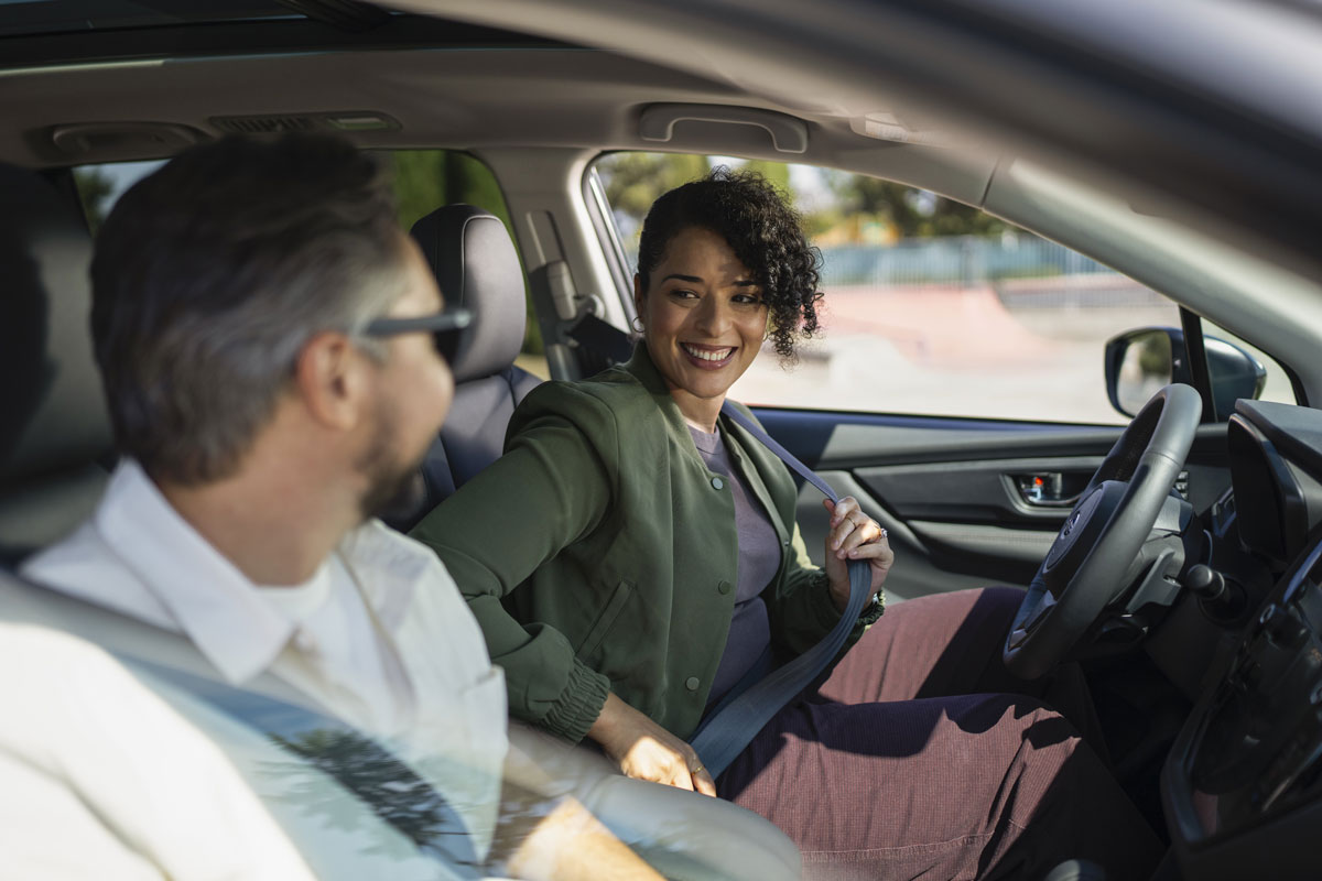 A woman in the driver's seat of a Subaru fastening her seatbelt and smiling at a man in the passenger seat.