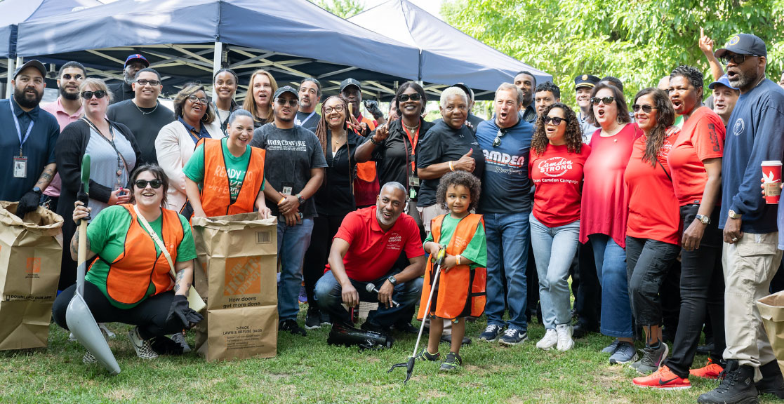 Group of smiling volunteers in vibrant clothing holding garden tools and bags and posing under canopies outdoors.