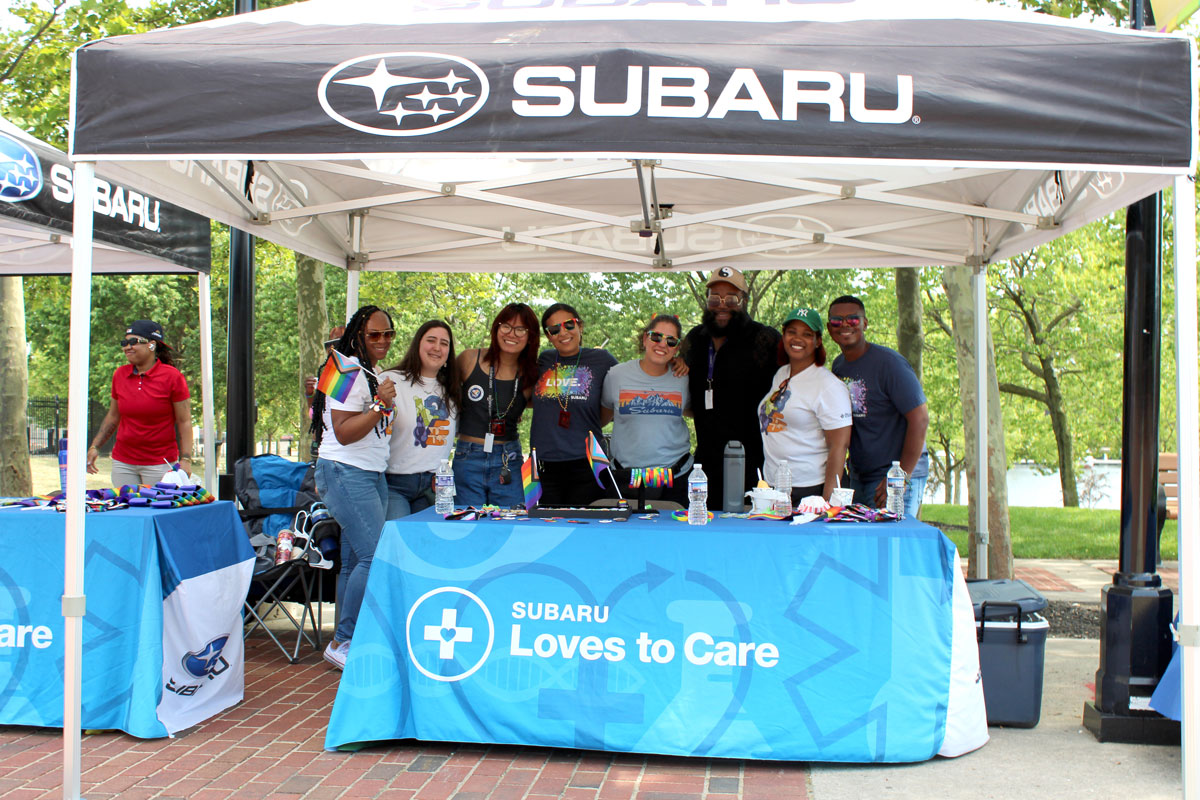 A group of people smiling under a tent at a Pride event, holding Pride flags and standing in front of a "Subaru Loves to Care" table.