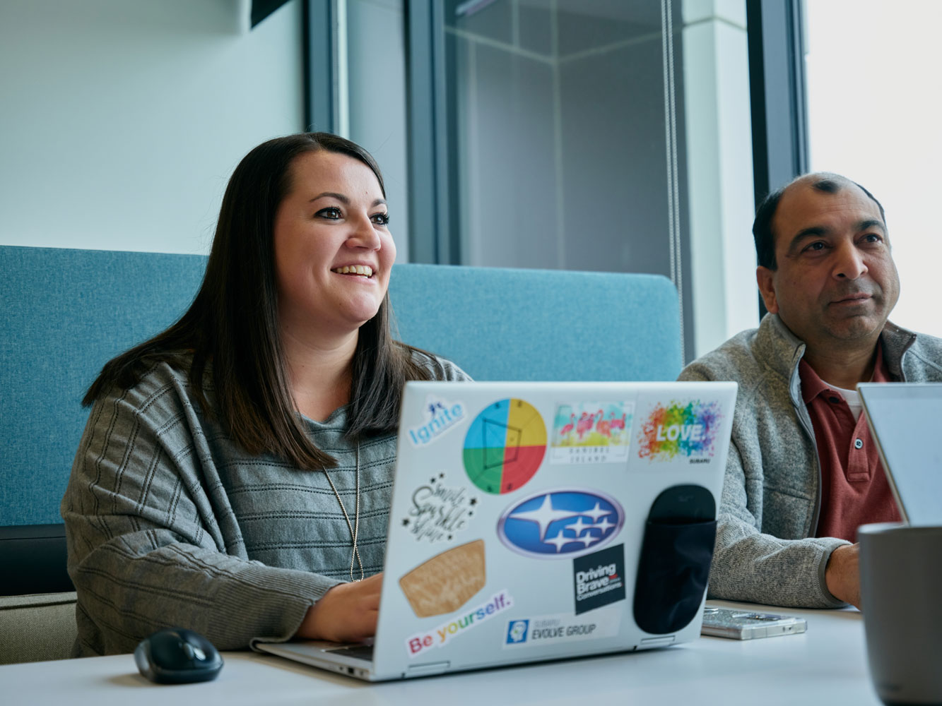 Smiling woman with long brown hair sits at a desk, using a laptop adorned with colorful stickers.