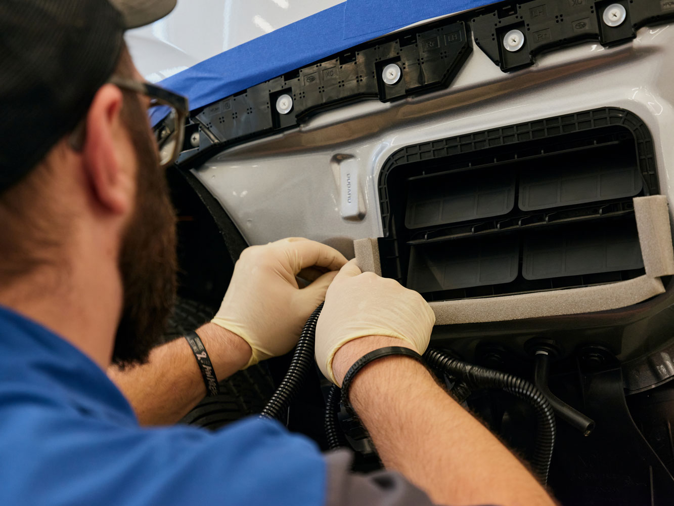 A man in a blue shirt and gloves works carefully on a car's front end, adjusting wiring.