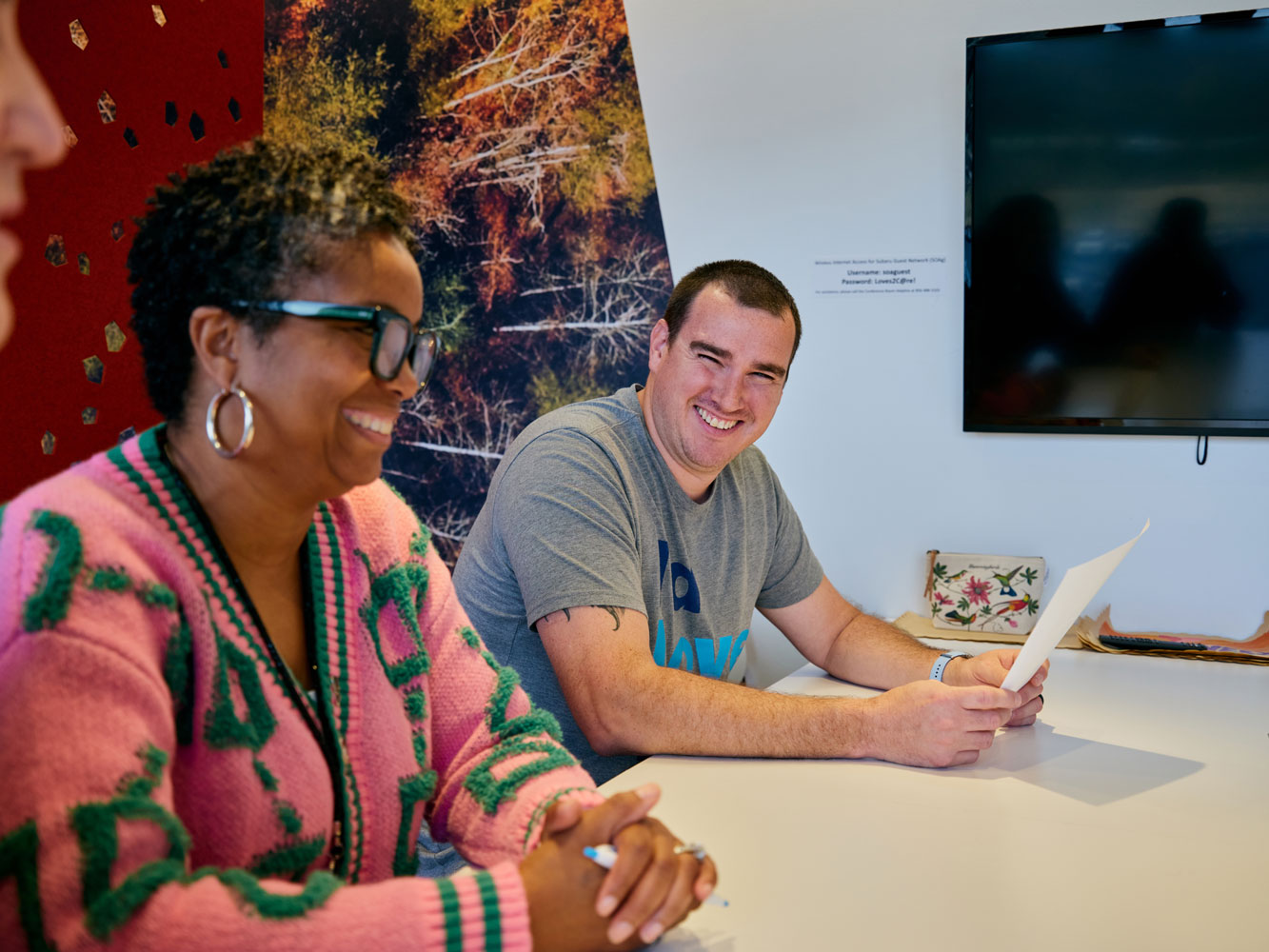 Two people sit at a table, smiling and engaged in conversation. The woman wears a pink and green sweater; the man wears a gray t-shirt.