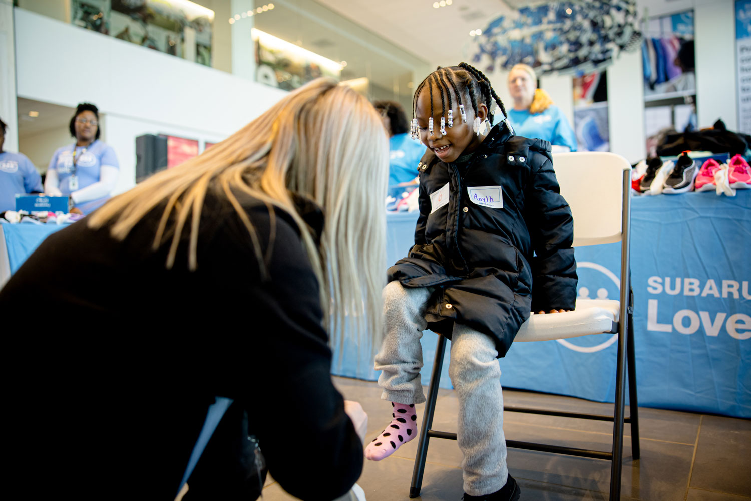 A child wearing a black coat and pink socks sits happily on a chair, as a blonde woman kneels to help. Event tables with shoes and people are in the background.