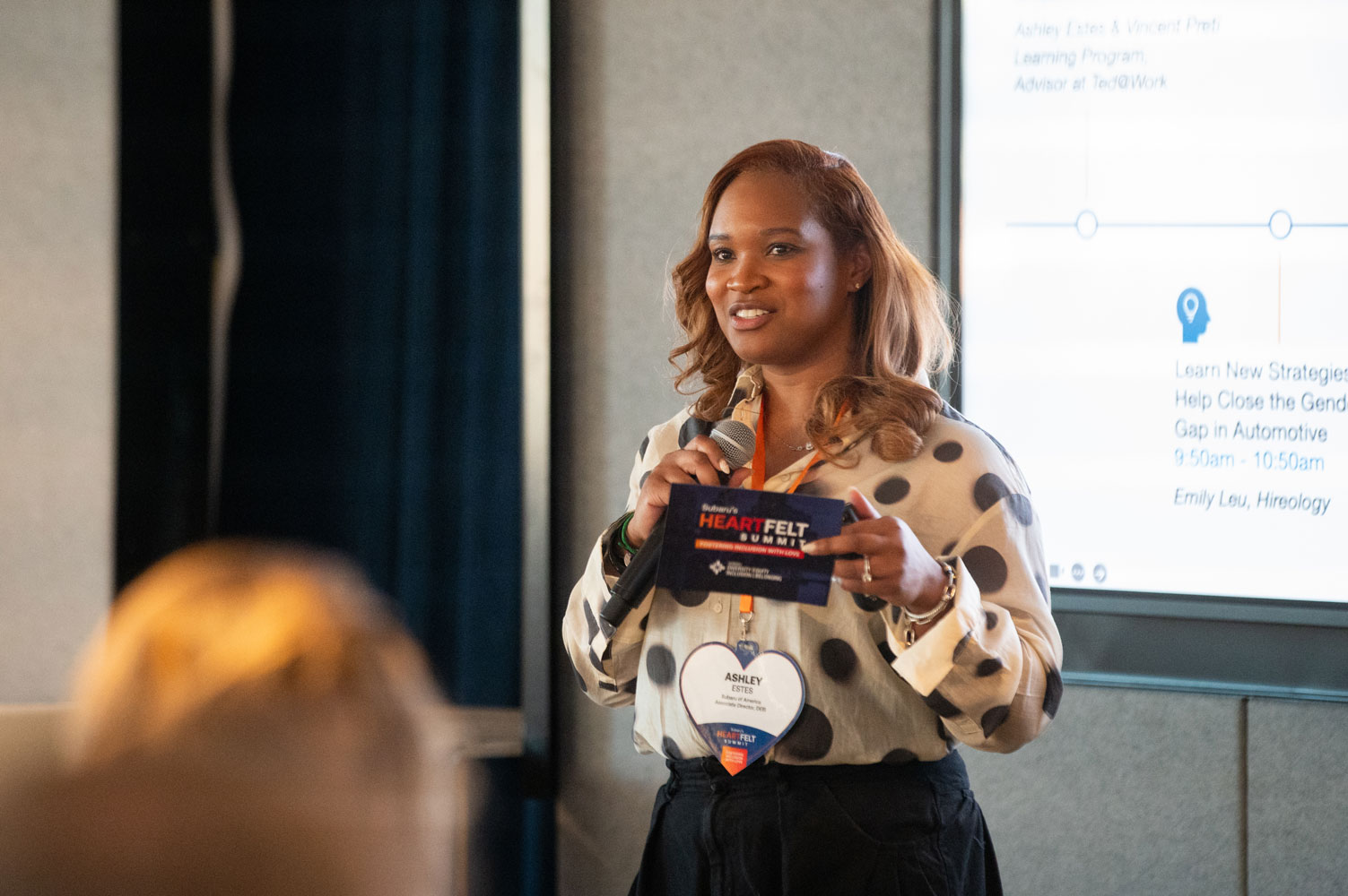 A woman wearing a polka dot blouse speaks confidently with a microphone, holding Subaru's Heartfelt Summit event materials.