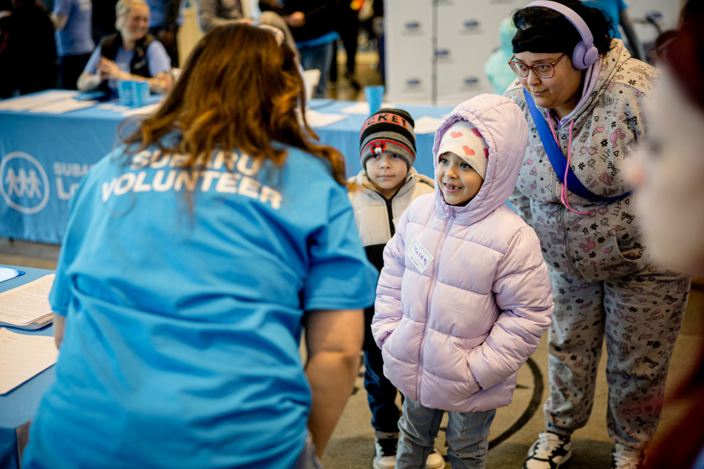 A volunteer in a blue shirt interacts with two children in warm jackets and a parent in a festive setting.