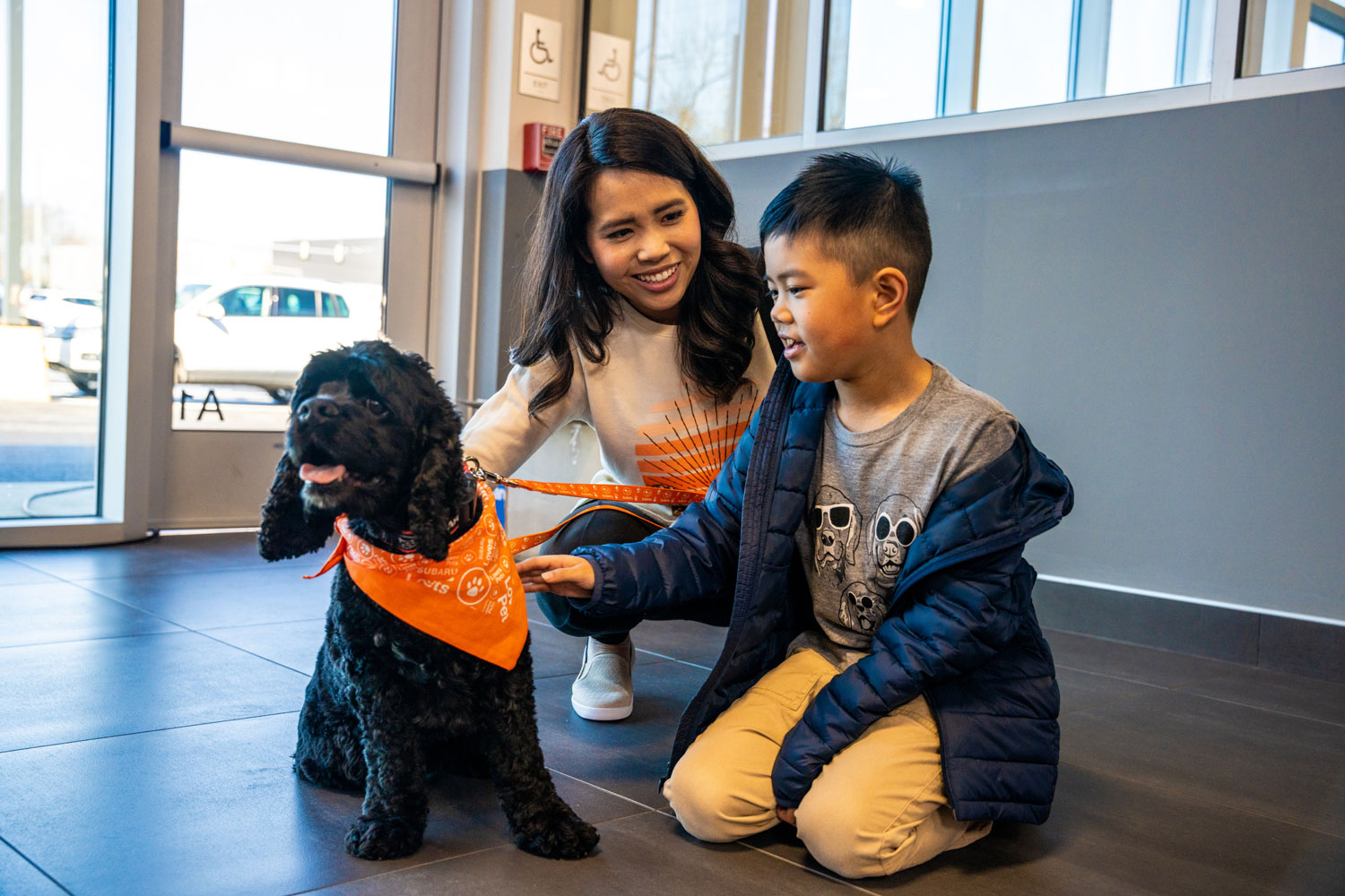 A woman and young boy smile and kneel beside a black dog wearing an orange bandana indoors.