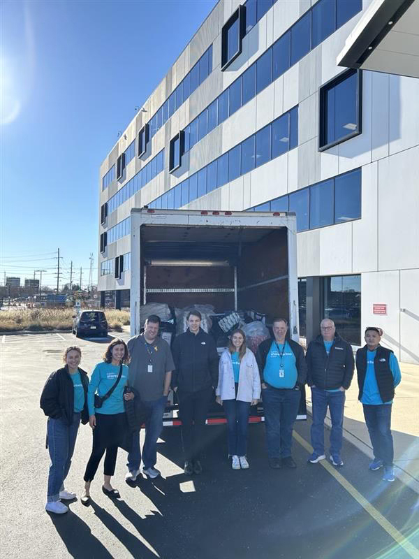 A group of eight smiling people stand in front of an open truck filled with bags, outside a modern office building under a clear blue sky.