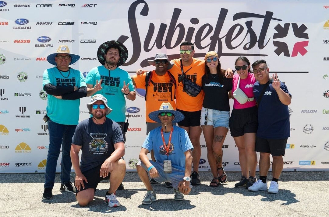 A group of seven smiling people in colorful T-shirts and hats pose in front of a "Subiefest" banner.