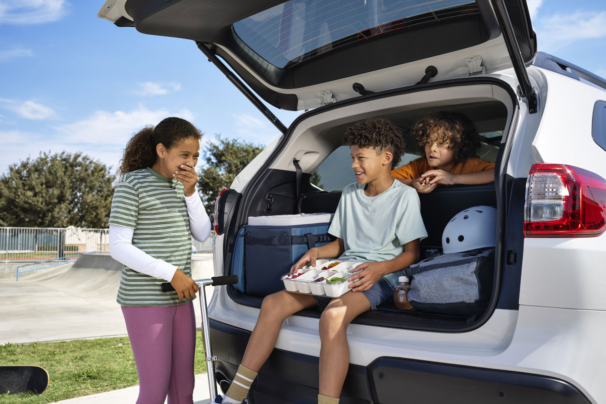 Three children enjoy a snack from the open trunk of a white Subaru Ascent SUV parked by a skate park. One stands with a scooter, smiling and laughing.