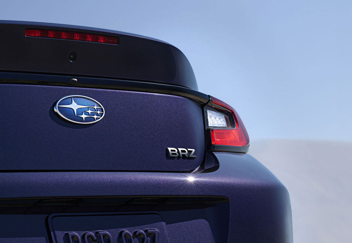 Rear view of a blue Subaru BRZ with logo and badge in focus. The car's sleek design and tail light contrast against a clear blue sky.