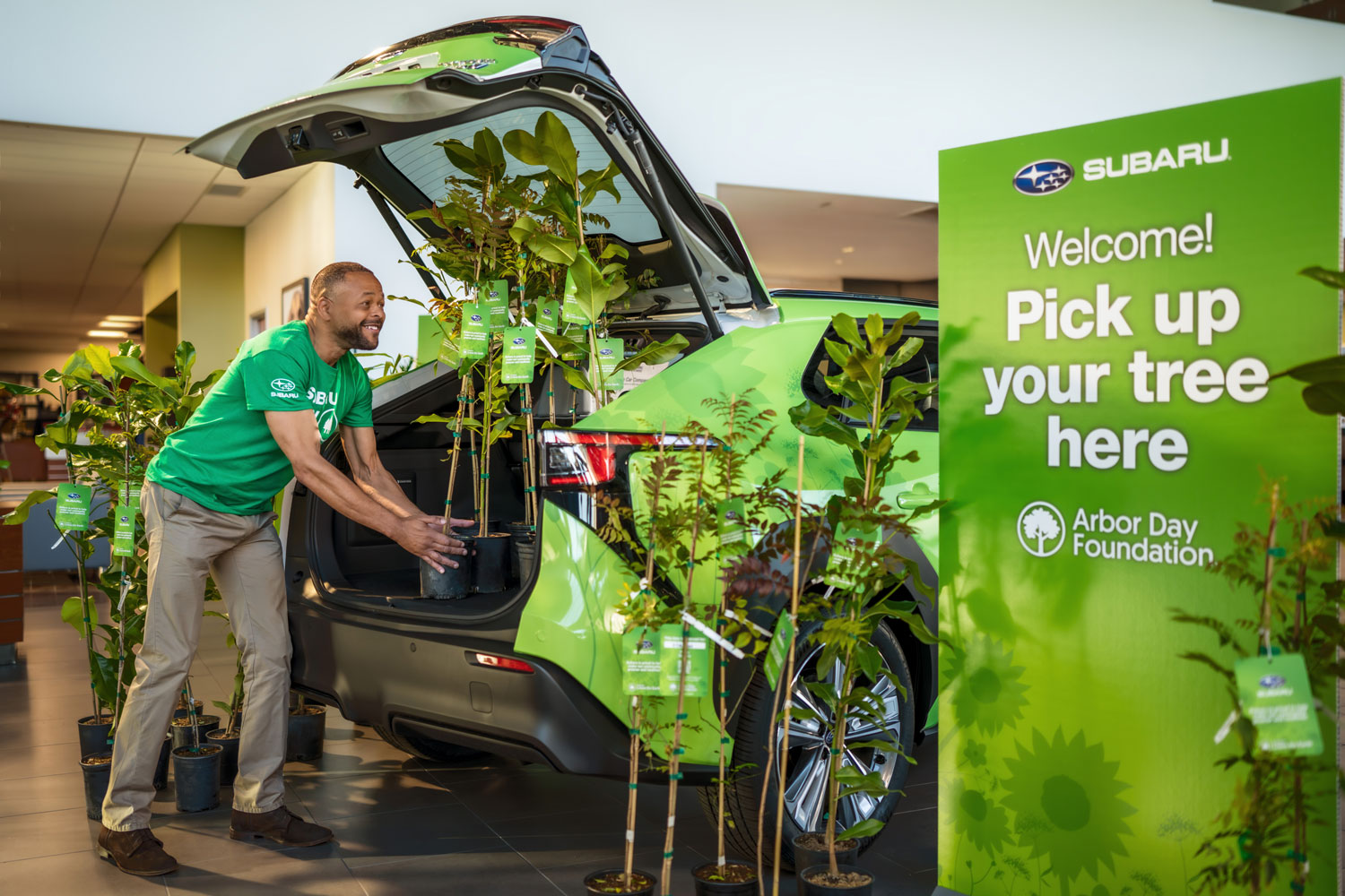 Man in a green t-shirt placing potted trees into a green car's trunk inside a showroom. A sign reads "Welcome! Pick up your tree here" from the Arbor Day Foundation.