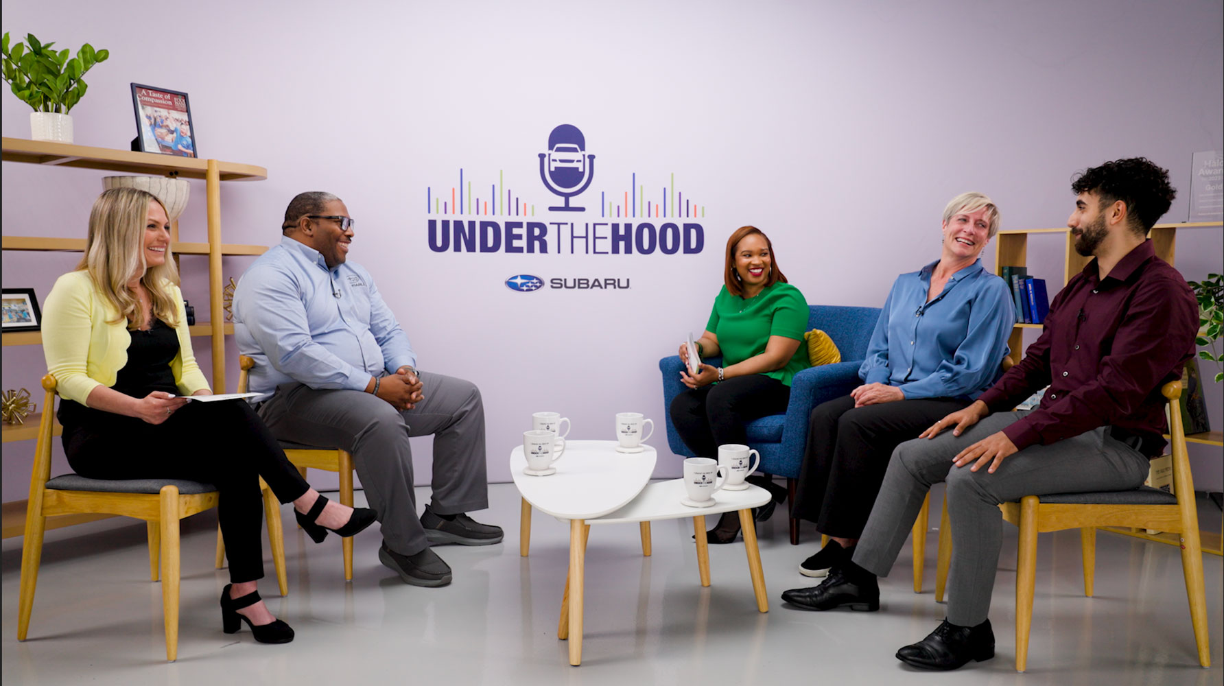 Five smiling people sit on modern chairs in a studio with