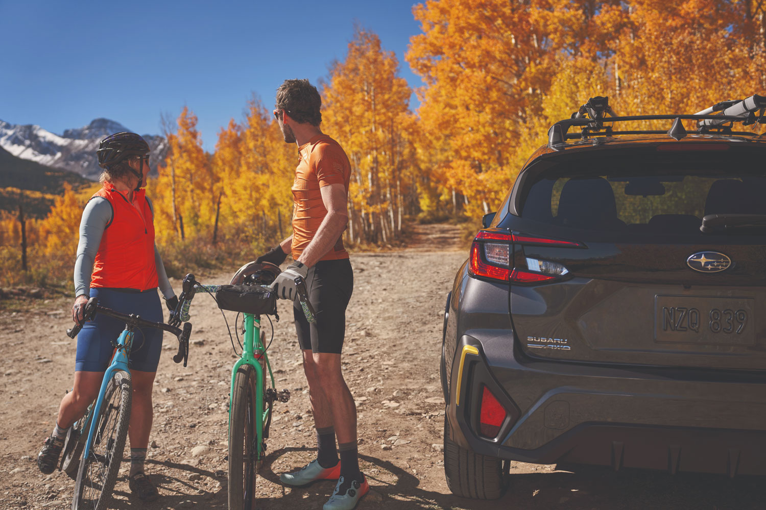 A cyclist in an orange shirt stands between a green bike and a parked gray Subaru SUV, looking at vibrant autumn trees.