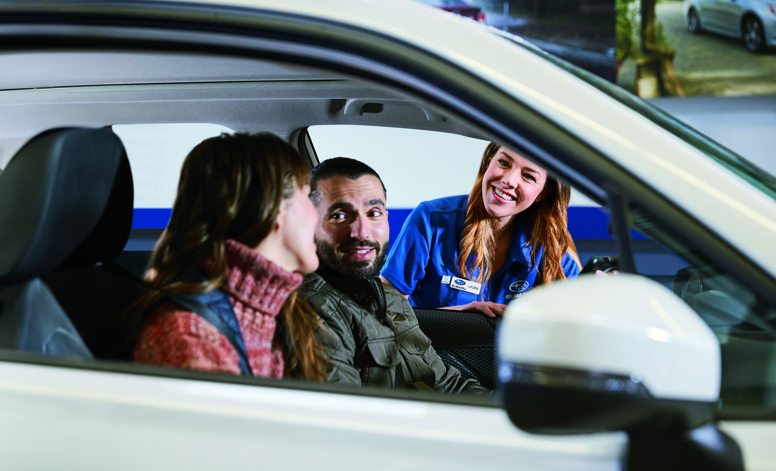 A smiling Subaru car service representative in a blue uniform speaks to a couple seated in a car.