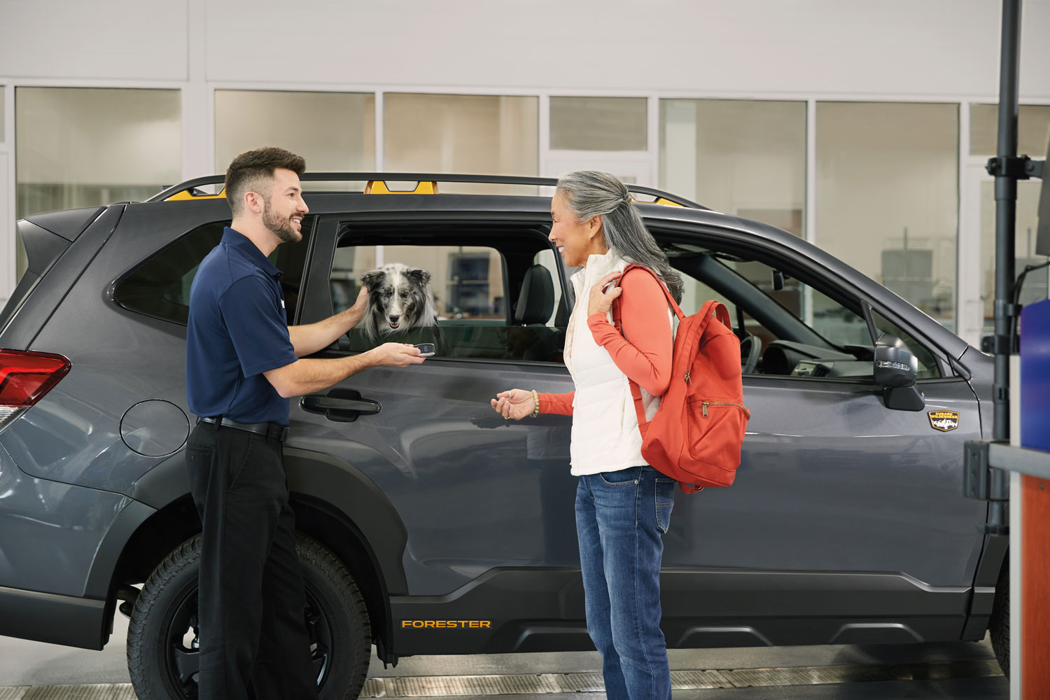 A smiling salesperson hands car keys to a happy woman standing by a gray Subaru SUV with a dog peering out the car window.