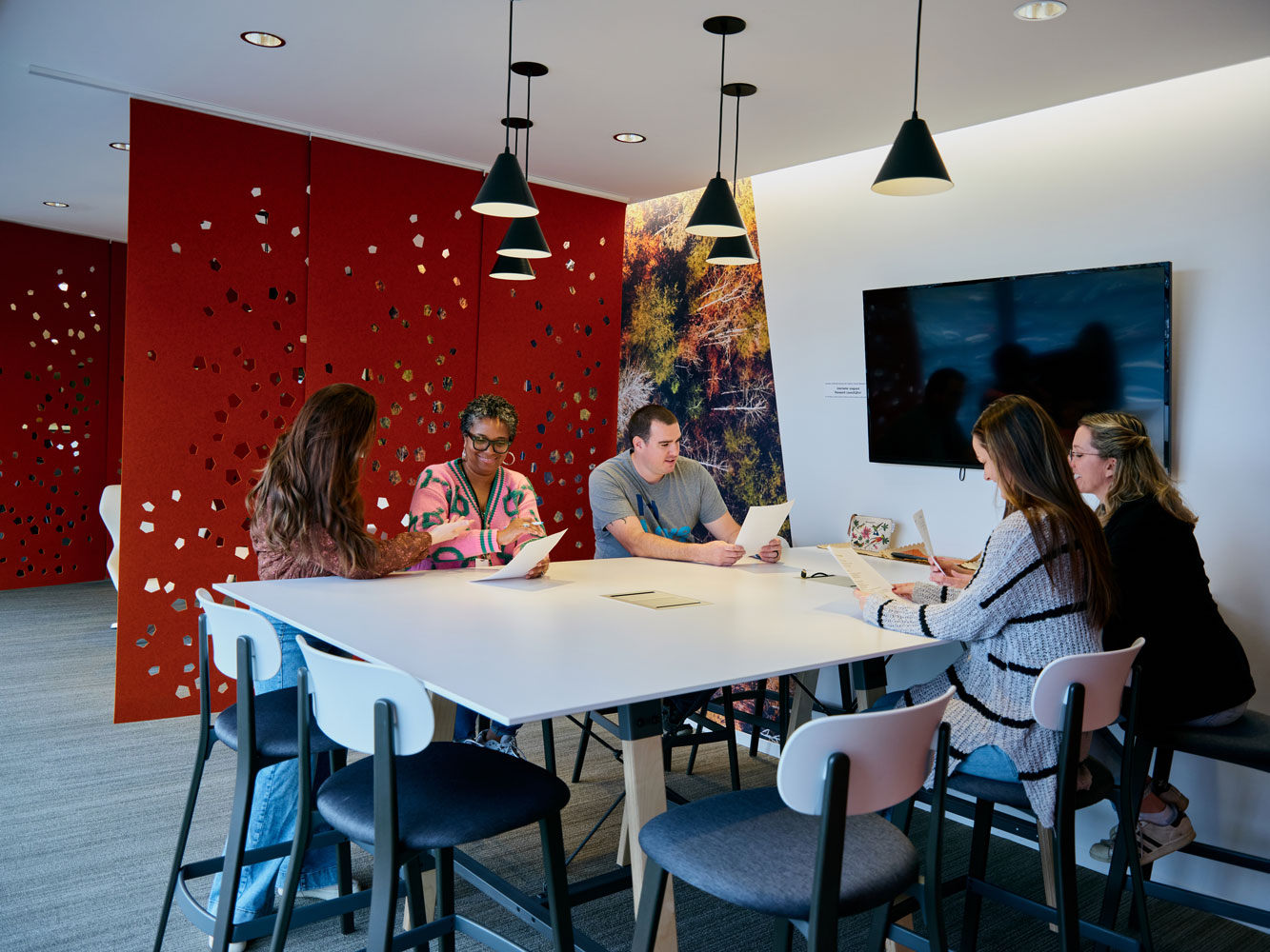 A diverse group of five people sit around a modern table, reviewing documents in a room with red and nature-themed walls.