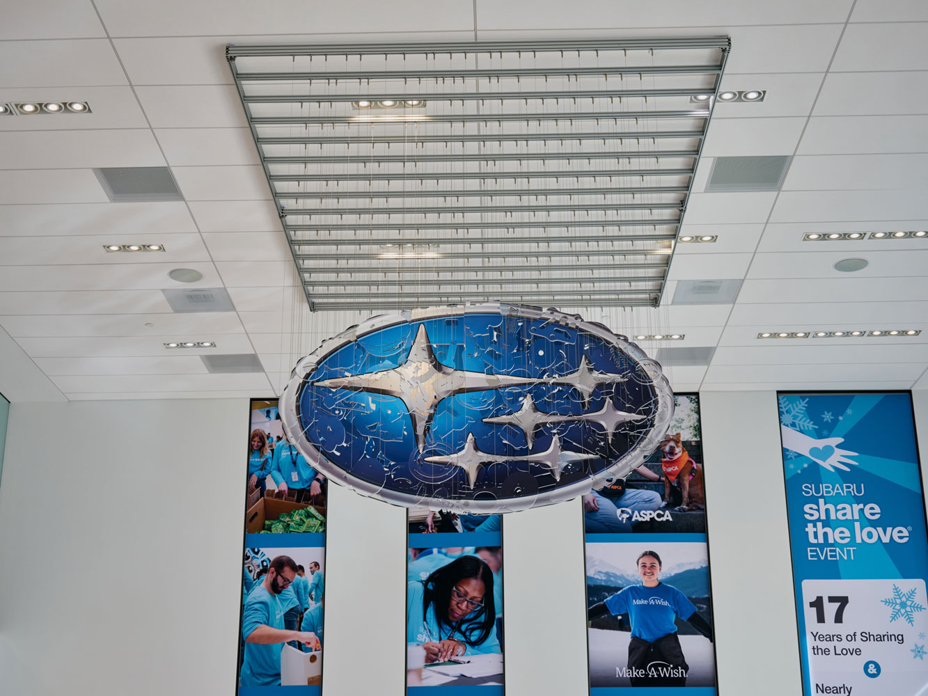 Hanging sculpture of the blue and silver Subaru emblem. Background features volunteer images from charities including Make-A-Wish and ASPCA.
