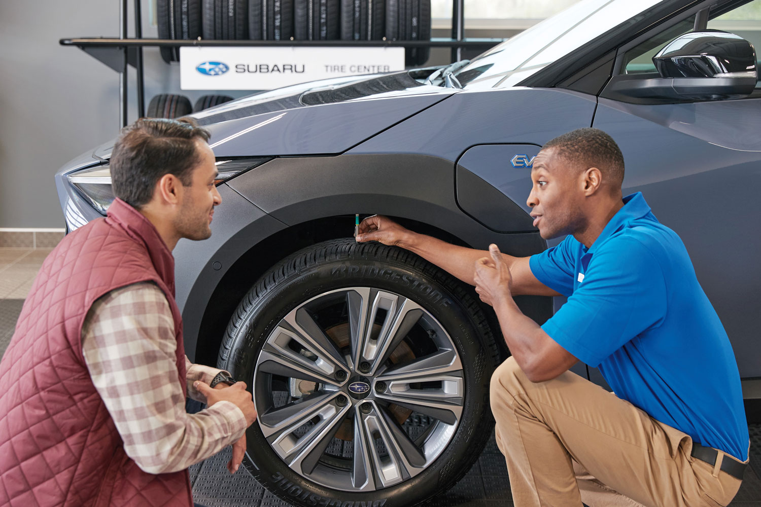 A man in a blue shirt demonstrates a tire gauge to another man beside a gray car at a Subaru tire center.