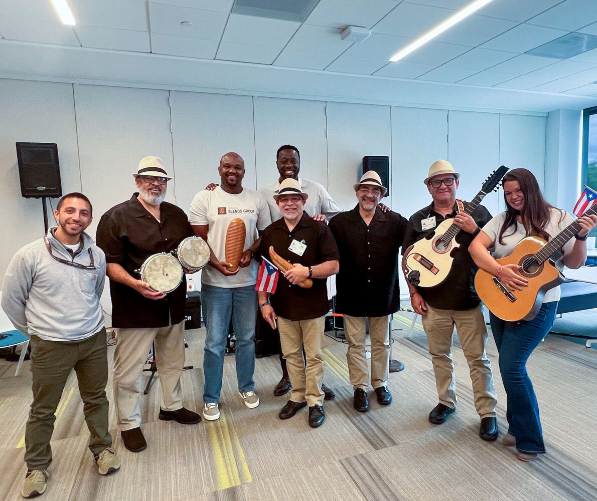 Group of seven people smiling in an indoor setting, holding musical instruments, including guitars and bongos.