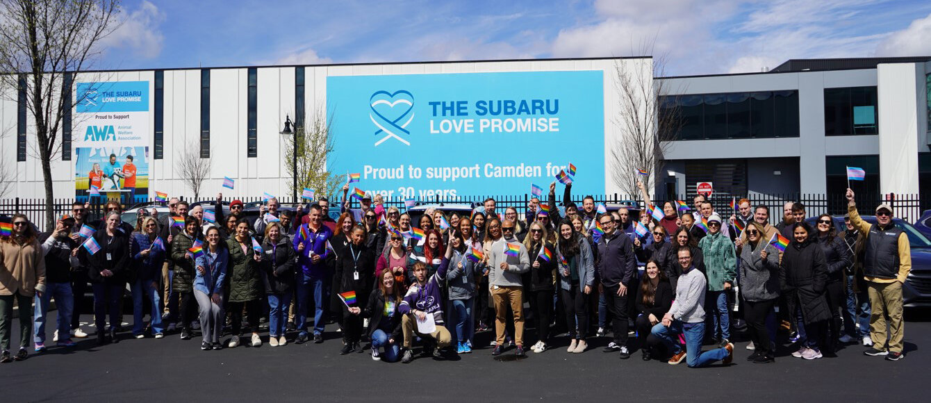 Group of people holding Pride flags and smiling outdoors in front of a large sign that reads "Proud to support Camden for over 30 years."