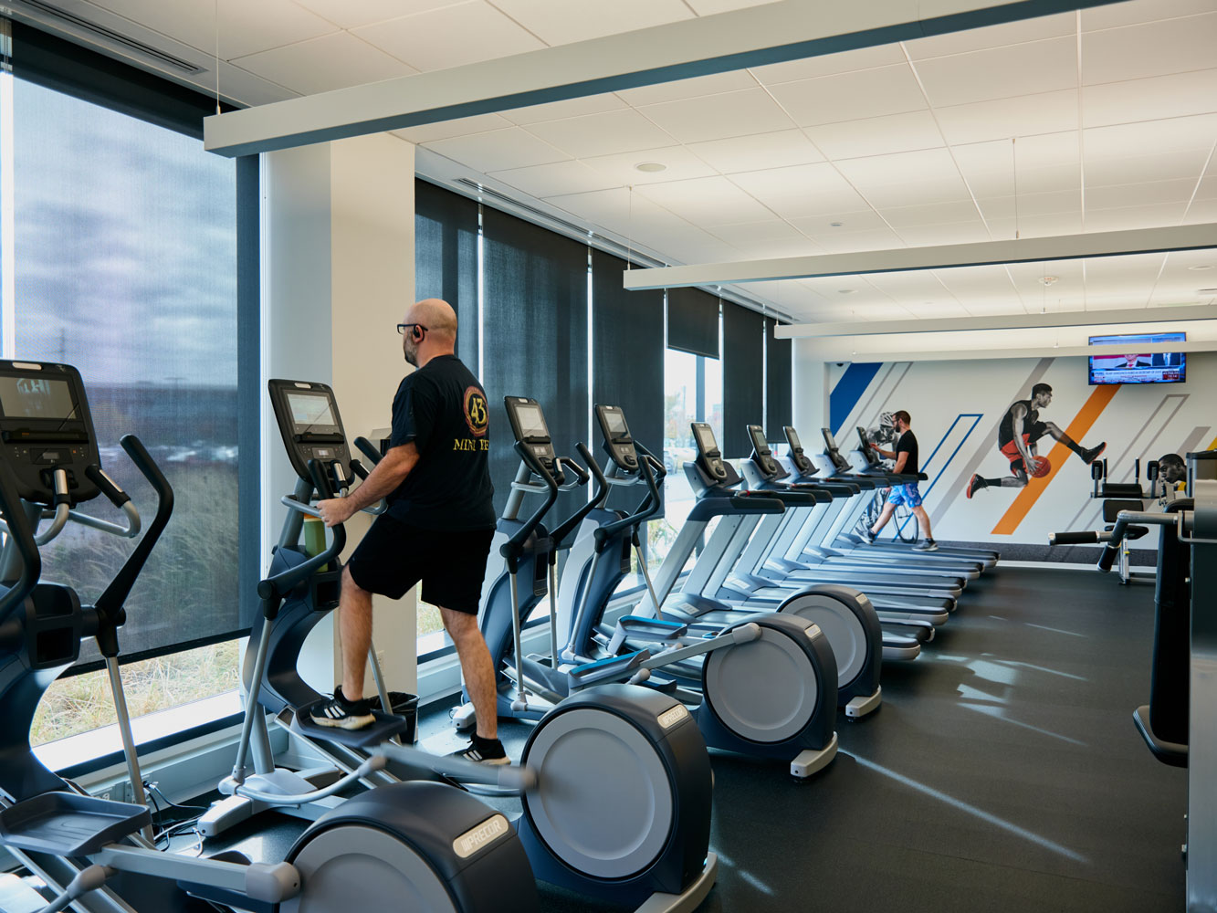 A man exercises on an elliptical machine at the gym, alongside large windows and several vacant machines.