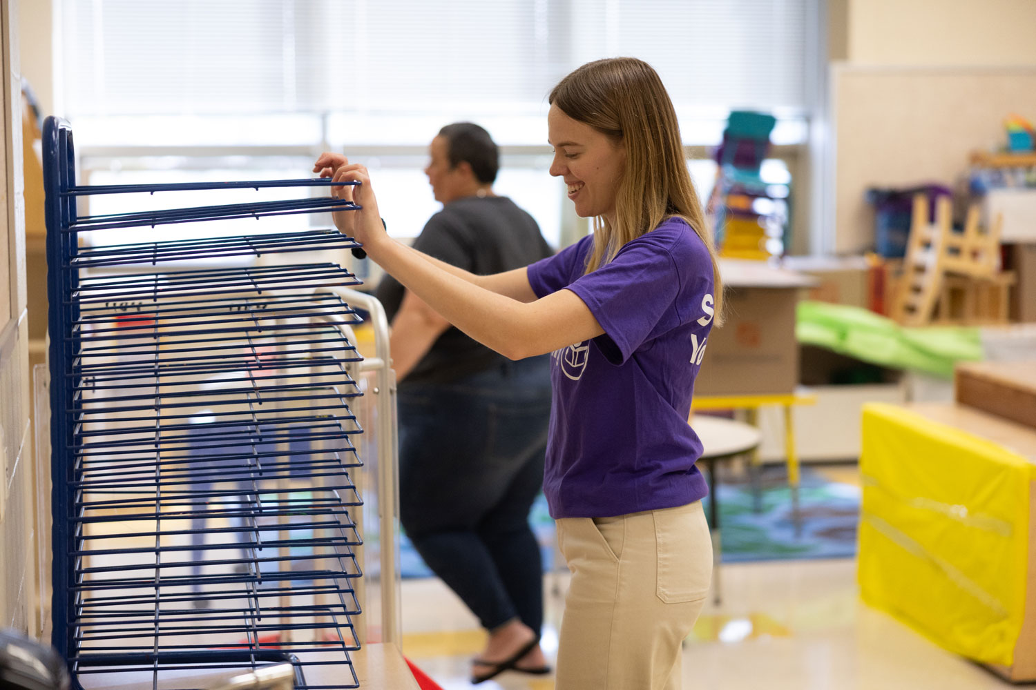 A woman in a purple shirt smiles while arranging a drying rack in a brightly lit classroom. Another person stands in the background.