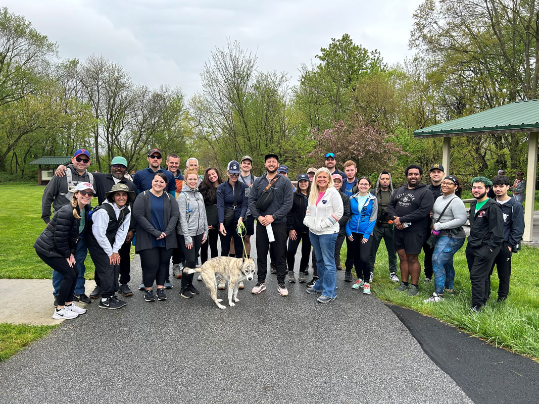 A diverse group of people and a dog smiling outdoors at a park, gathered on a pathway with trees and greenery in the background.