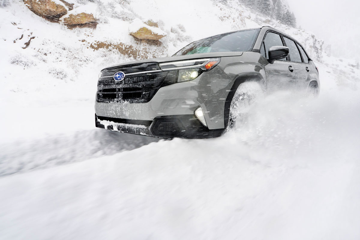 A gray Subaru Forester SUV drives through a snowy landscape with snow-covered rocks in the background, kicking up snow around its wheels.