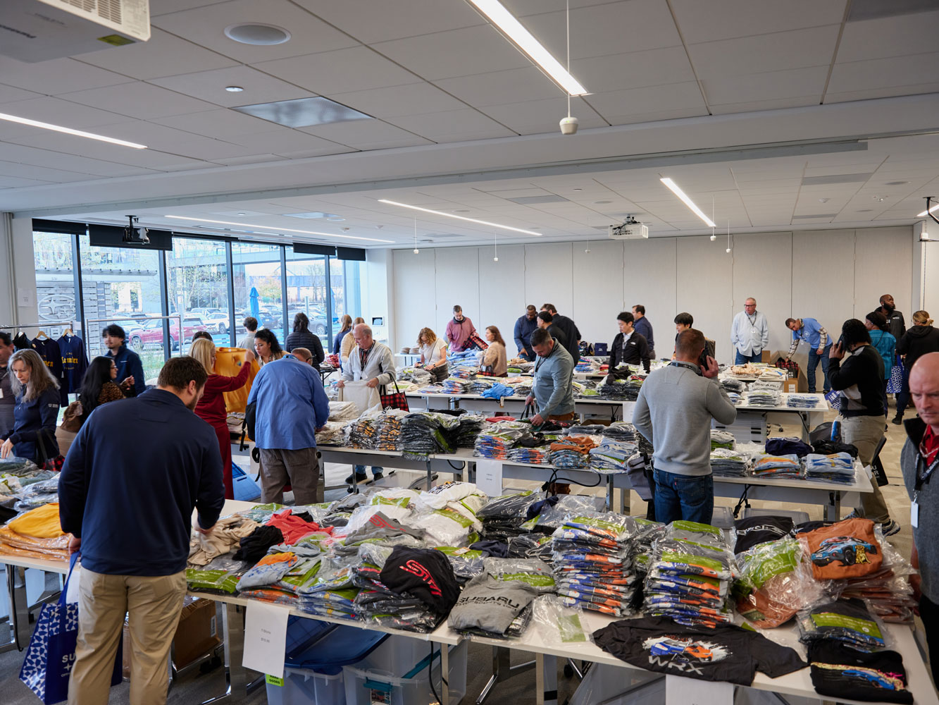 A group of people in a large room sorts through neatly organized Subaru t-shirts on tables.