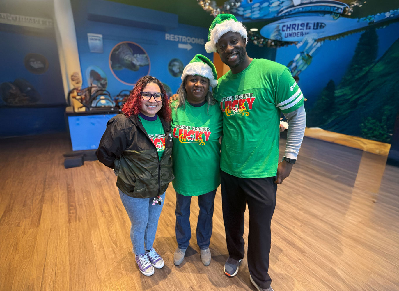 Three people smile in a holiday-themed setting, wearing green shirts with