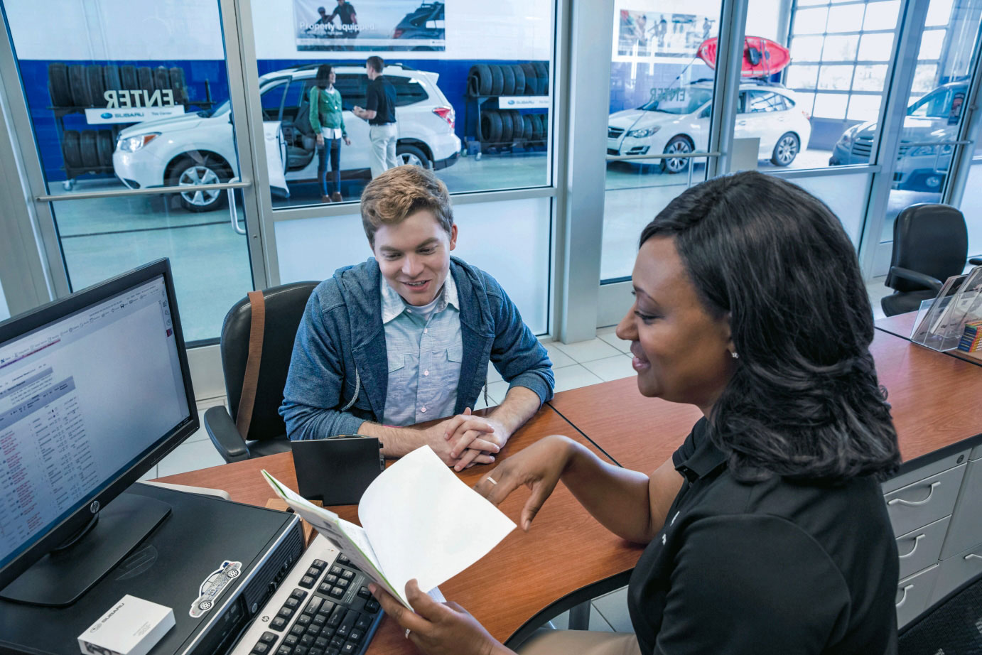 Saleswoman and customer smiling while discussing a document at a car dealership. Background shows a showroom with several cars.