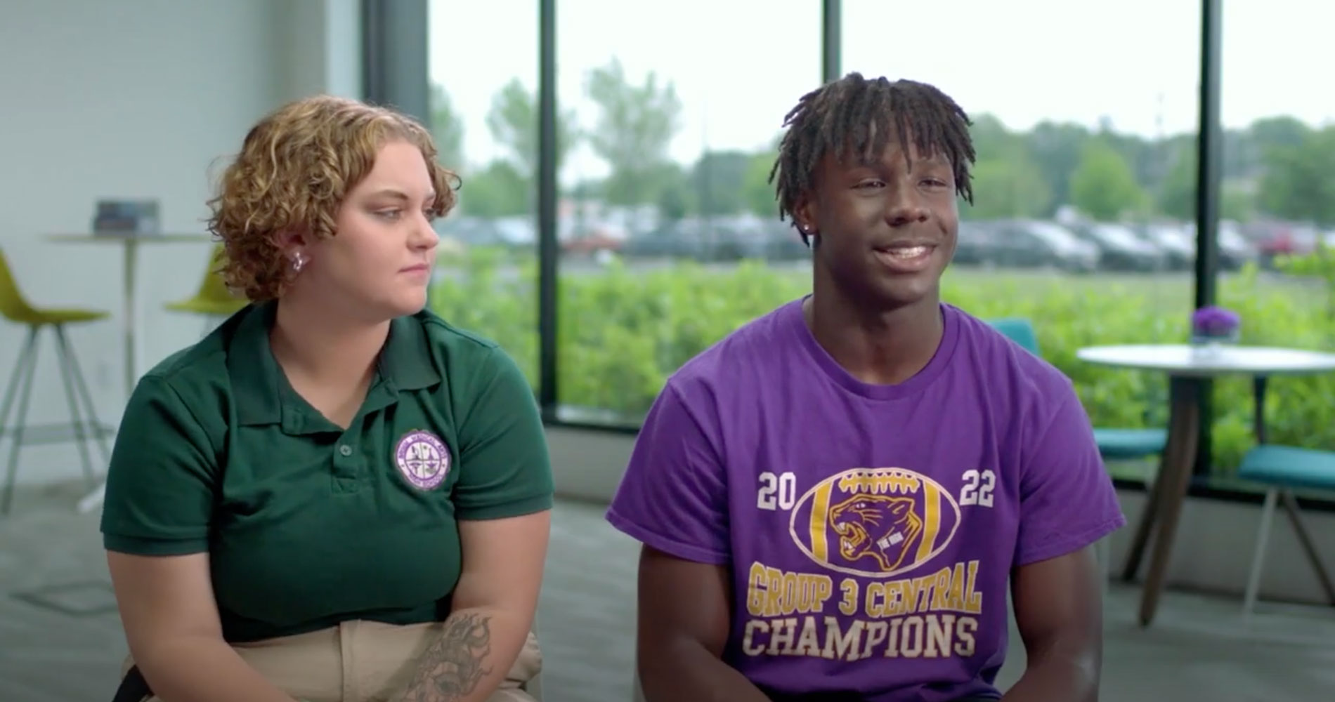 Two people sit indoors by a large window. The person on the left wears a green polo shirt with a logo, looking at the smiling person in a purple "2022 Group 2 Central Champions" shirt. The background is bright, with a table and chairs visible.