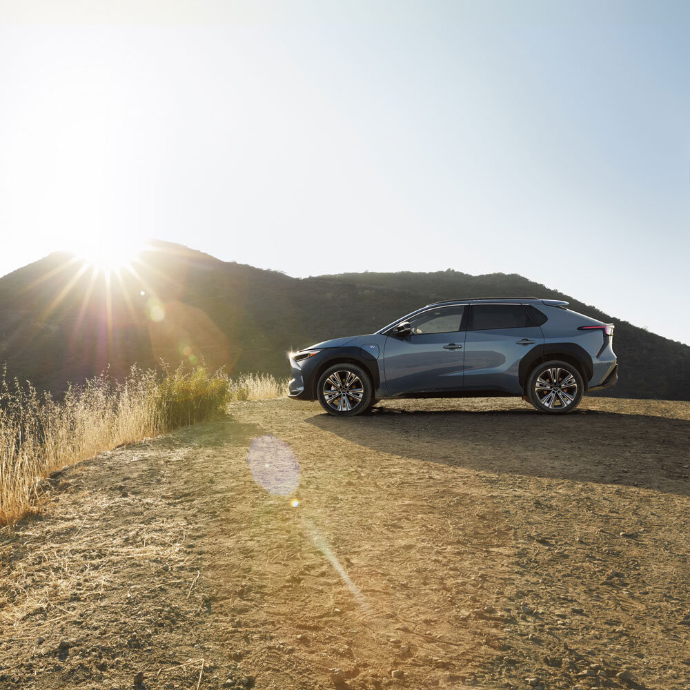 A sleek, blue Subaru SUV parked on a sunlit hilltop with dry grass, against a backdrop of mountains.