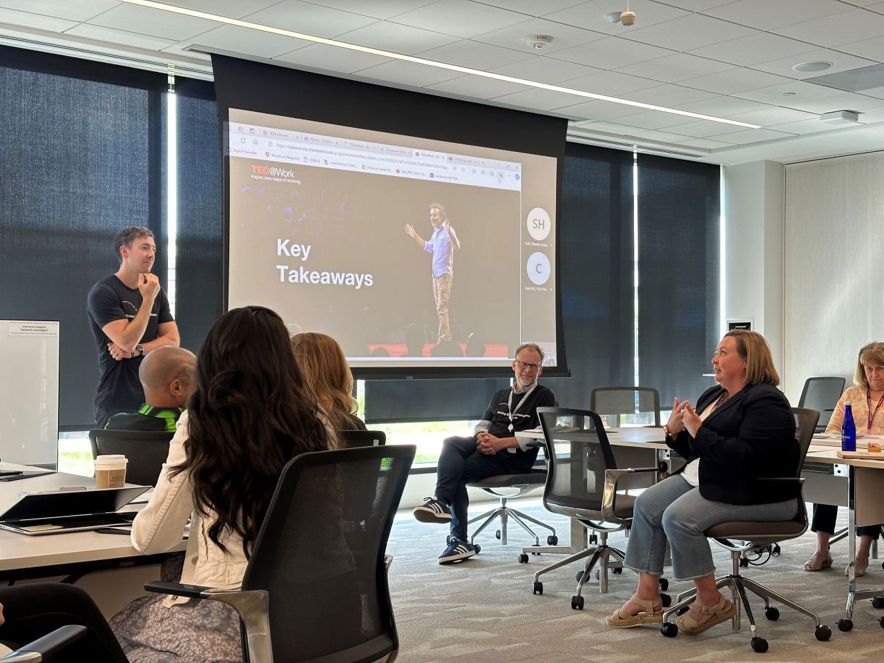 A group of people in a conference room watch a TED@Work presentation featuring a "Key Takeaways" title on a screen.