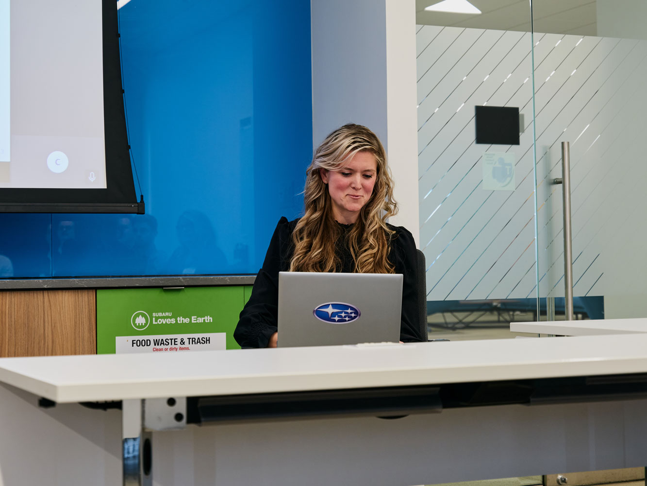 A woman sits at a conference table using a laptop, with a thoughtful expression. Behind her, there is signage about waste management and a projection screen.