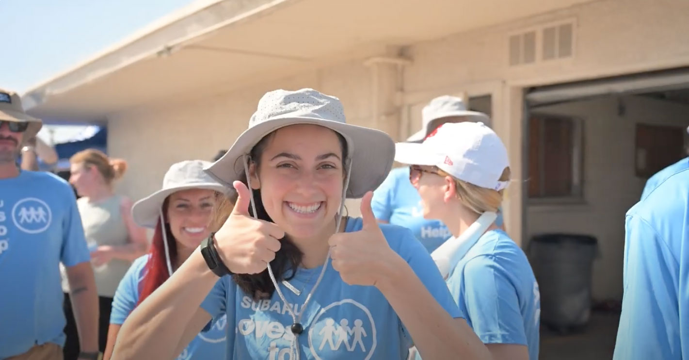 Smiling woman gives two thumbs up, wearing a sun hat and blue Subaru event T-shirt, surrounded by others in matching shirts.