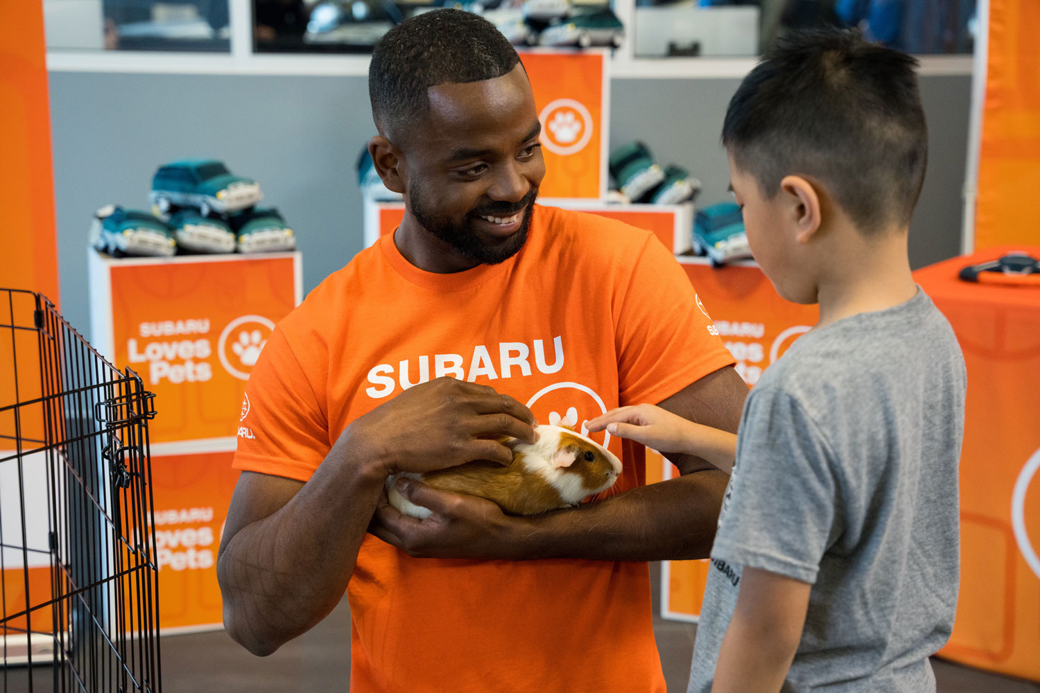 A man in an orange "Subaru Loves Pets" shirt holds a guinea pig, smiling at a young boy petting it.