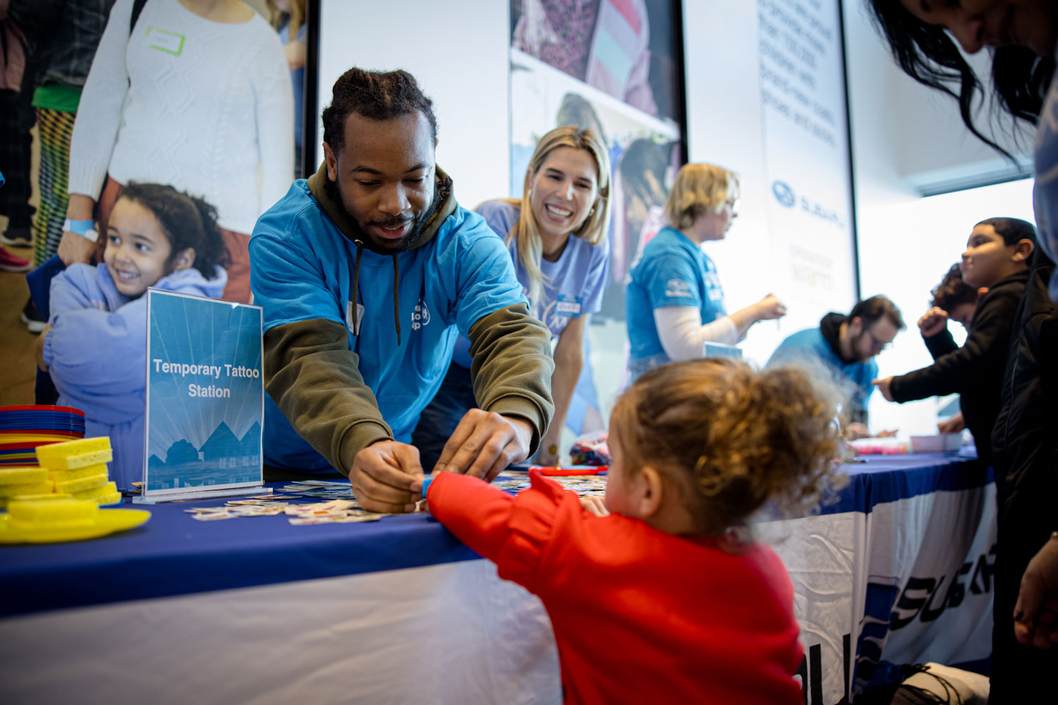 A man at a temporary tattoo station helps a child in a red sweater. Smiling volunteers in blue shirts assist other children nearby.