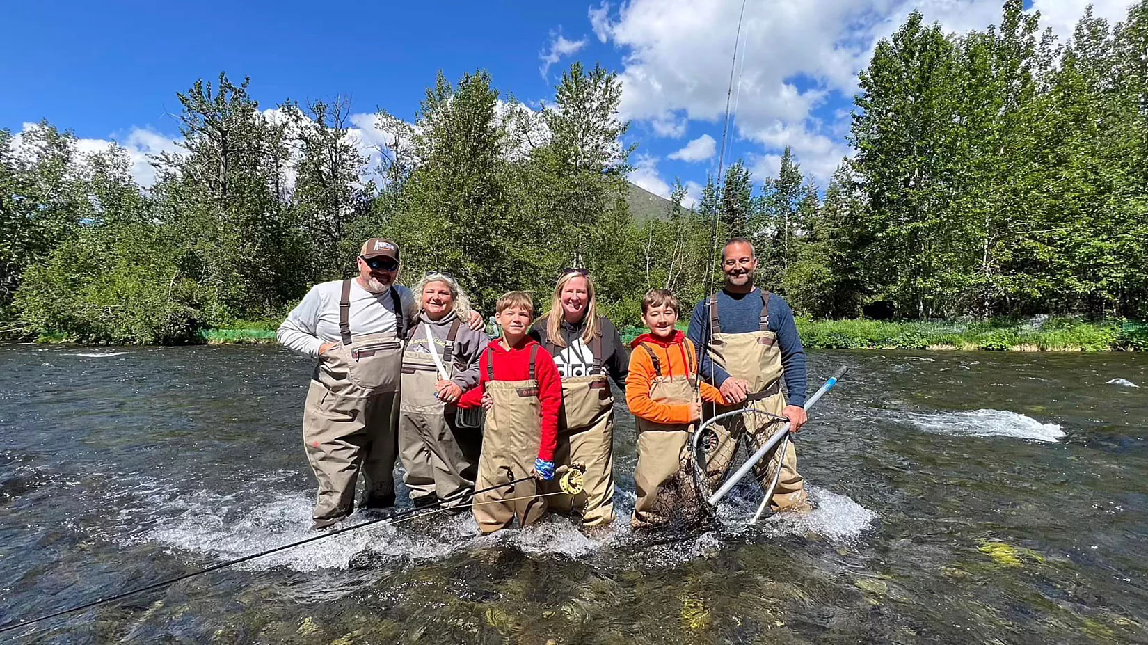 A group of six people in fishing waders stands in a clear, shallow river, holding fishing rods and nets. They're smiling under a bright blue sky with trees in the background.