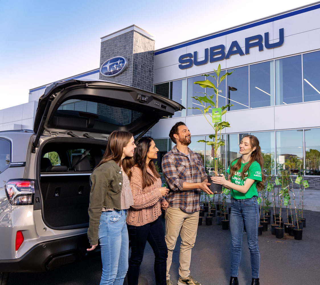 A group of four people, smiling, stand beside an open SUV trunk in front of a Subaru building. One person hands over a potted plant.