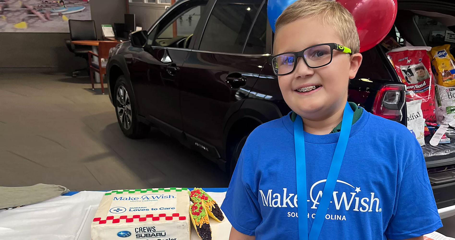 Smiling boy wearing glasses and a Make-A-Wish T-shirt stands beside a cake decorated with tacos. A car and balloons are in the background.