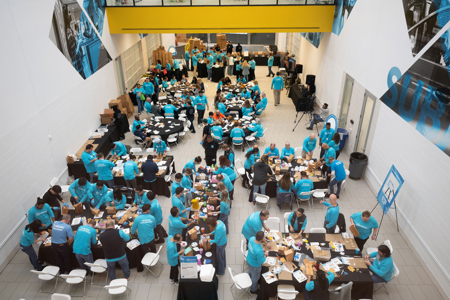 A large group of people in teal shirts gather in a bright, spacious room with white walls. They are seated at round tables, actively working on projects and socializing.