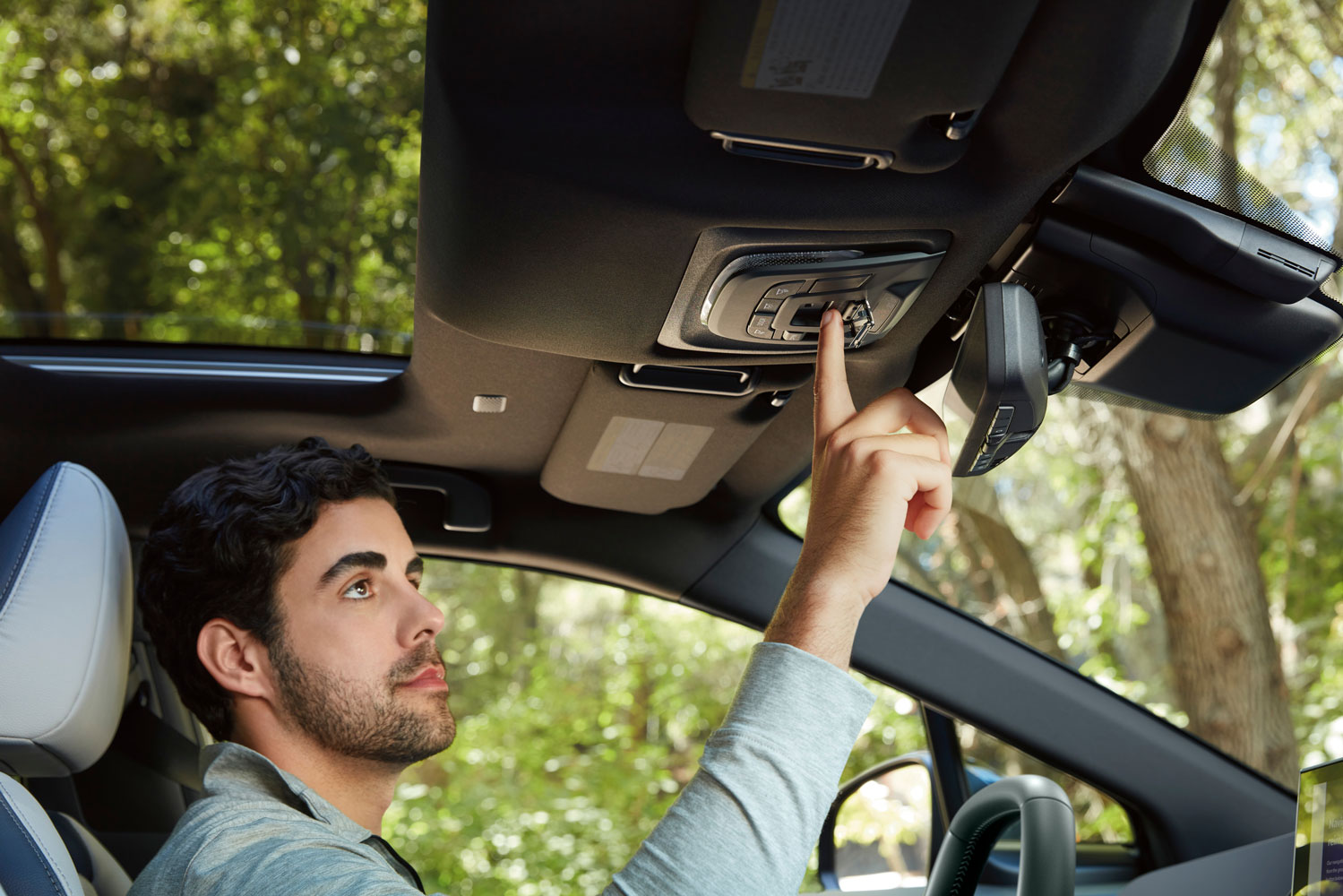Man in a car interior adjusts a control on the ceiling. He's focused, surrounded by lush greenery visible through the windows.