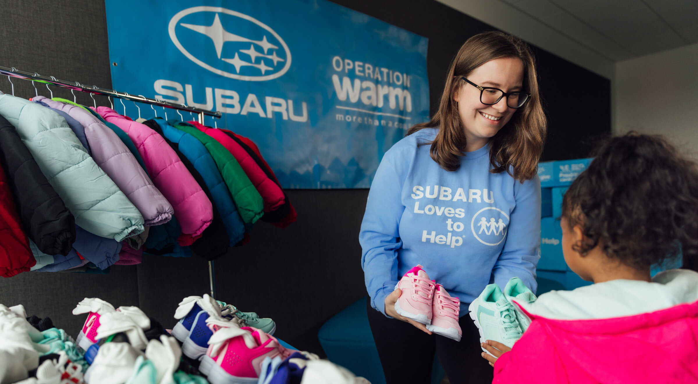 A smiling woman in a blue "Subaru Loves to Help" shirt hands pink shoes to a young girl in a bright pink coat. Various colorful coats and shoes are visible.