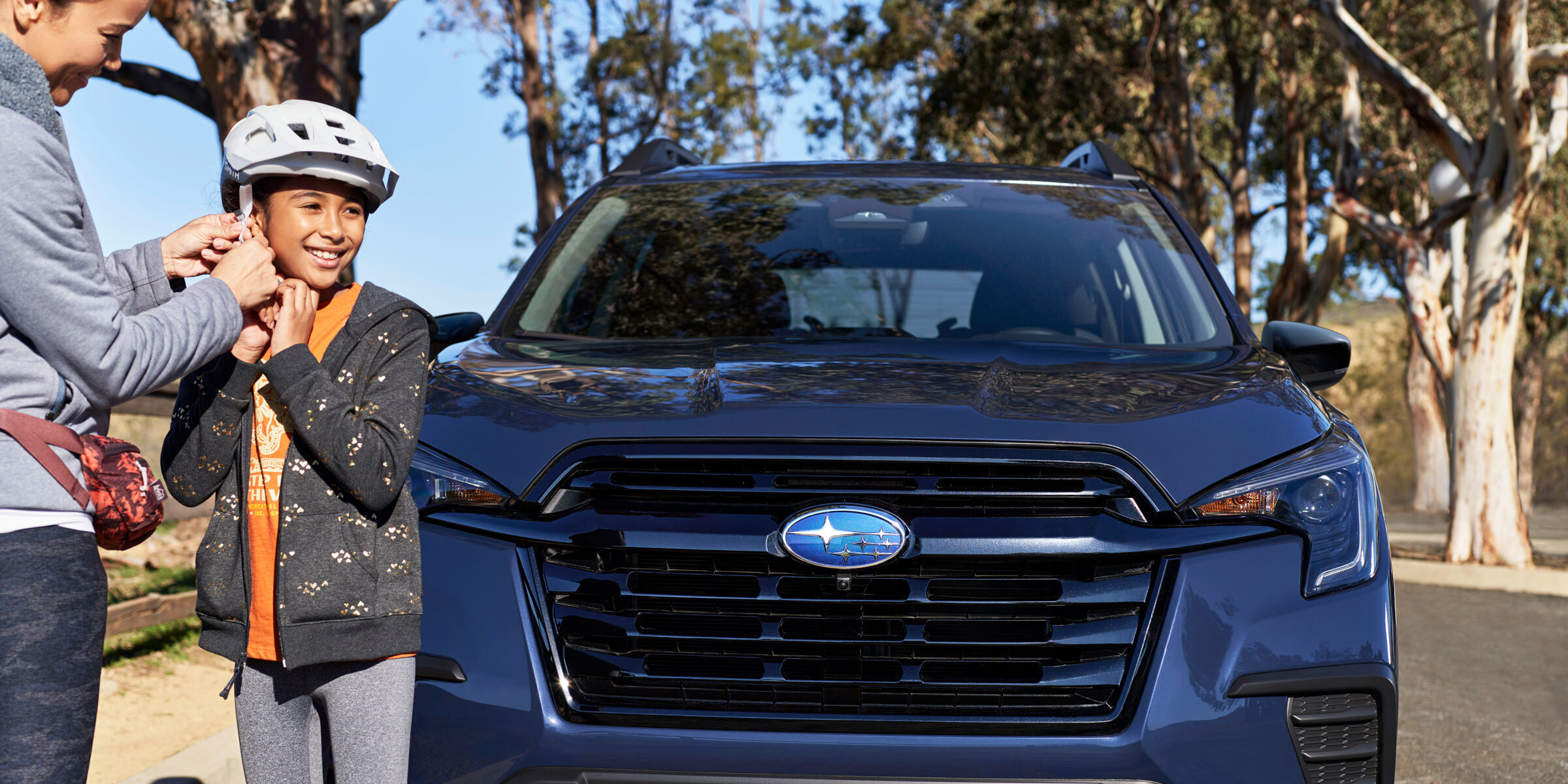 A woman fastens a white helmet on a smiling girl next to a dark blue Subaru parked on a sunny road.